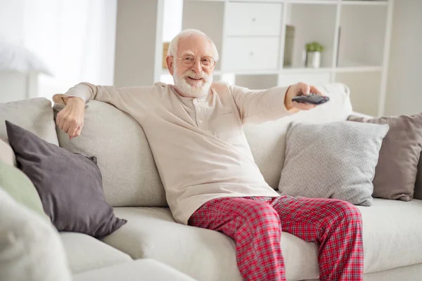 Фото літнього чоловіка щаслива позитивна посмішка сидячий диван будинок весело дивитися телевізійний перемикач канал пульт дистанційного керування — стокове фото
