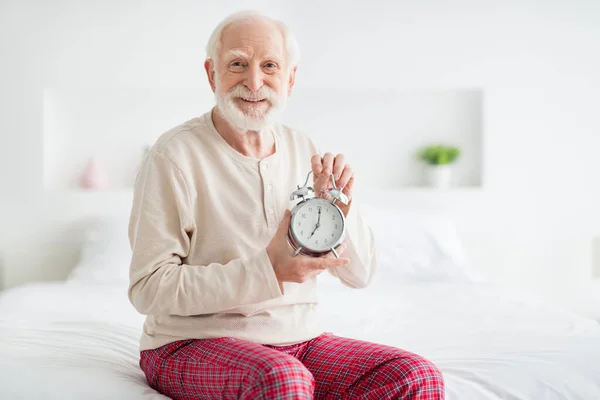 Photo of elderly man pensioner sit on bad home happy positive smile hold clock alarm timer wear pajamas