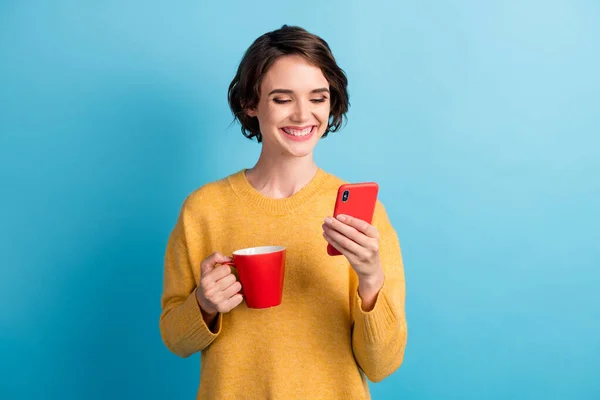 Foto de mujer dentada radiante vestida casual suéter amarillo bebiendo café buscando teléfono aislado color azul fondo — Foto de Stock