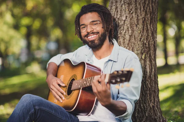 Foto portret van de man in glazen glimlachend zittend in groen park onder boom gitaar spelend in casual kleding — Stockfoto