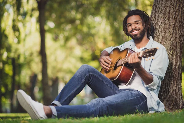 Portret van een aantrekkelijke vrolijke man zittend op groen gras spelen gitaar zingen romantische hit zonnige dag buiten — Stockfoto