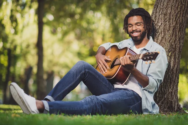 Full length foto van afro-Amerikaanse jongeman zitten gras dragen bril spelen gitaar buiten in park buiten — Stockfoto