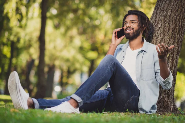 Pełna długość zdjęcie brunetka zrelaksowany ciemna skóra człowiek rozmawiać telefon siedzieć trawa w parku dobry nastrój na zewnątrz w plenerze — Zdjęcie stockowe