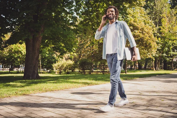 Full längd foto av mörk hud charmig brunett kille hålla laptop telefon tala promenad i park helg utomhus — Stockfoto
