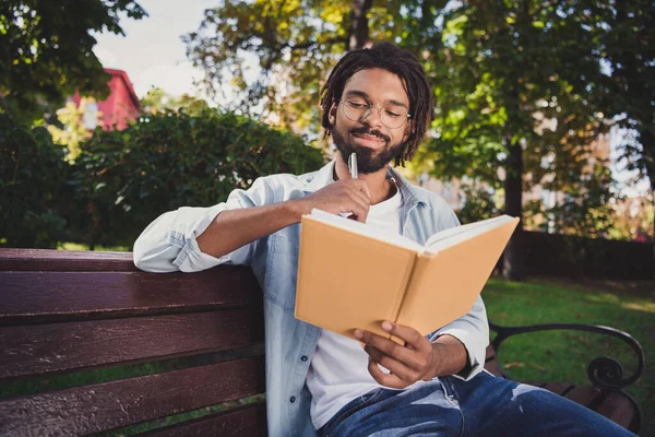 Foto van minded slimme jonge donkere huid man houden pen kin look lees notebook buiten in het park op bank buiten — Stockfoto