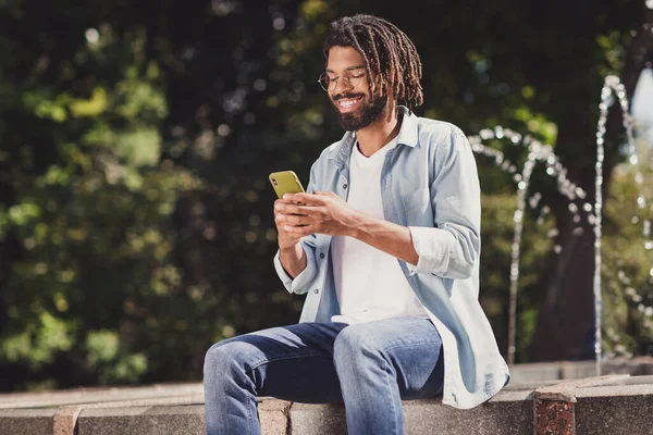 Retrato de chico alegre atractivo sentado cerca del agua en el aire libre utilizando el dispositivo de chat de navegación medios urbanos al aire libre — Foto de Stock