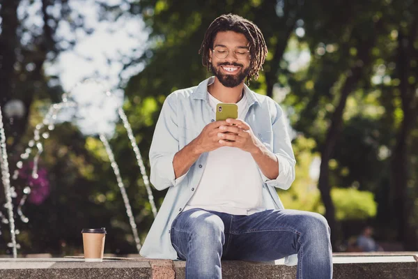 照片上快乐的年轻黑人男子手持电话，阅读短信，戴着眼镜坐在外面的公园里 — 图库照片