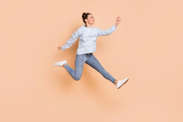 Foto de perfil de tamaño completo de optimista señora morena agradable salto captura desgaste suéter azul jeans zapatillas aisladas sobre fondo beige —  Fotos de Stock