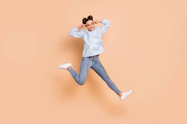 Foto de perfil de tamaño completo de optimista agradable dama morena salto mostrar v-signo de desgaste azul suéter jeans zapatillas aisladas sobre fondo beige —  Fotos de Stock