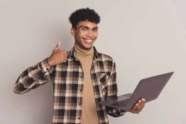 Foto retrato de un niño joven con camisa a cuadros que muestra un signo de pulgar hacia arriba sonriendo manteniendo la computadora aislada sobre un fondo de color gris —  Fotos de Stock