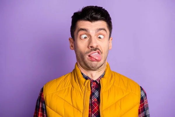 Foto de divertido joven infantil usar chaqueta amarilla sin mangas que muestra la lengua buscando nariz aislado color violeta fondo —  Fotos de Stock