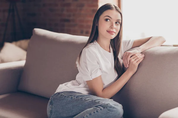 사려깊고 상냥 한 젊은 아가씨의 사진 흰색 티셔츠를 입고 소파에 앉아 미소를 짓고 실내 방에서 휴식을 취하는 모습 — 스톡 사진