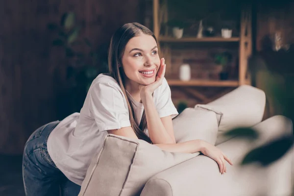Фото вдумчивой блестящей юной леди в белой футболке, сидящей на диване, улыбающейся, отдыхающей, глядя в окно в комнатах — стоковое фото