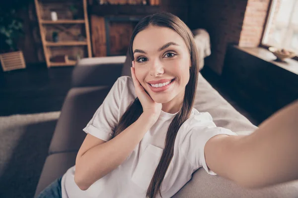 Foto van dromerige mooie jonge vrouw wit t-shirt zit bank arm wang spijkeren selfie glimlachen binnen appartement kamer — Stockfoto