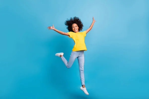 Full kropp foto av glad afro amerikansk flicka höja händerna hoppa bära gul t-shirt jeans isolerad på blå färg bakgrund — Stockfoto