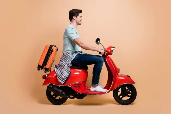 Profil boční pohled portrét pěkné veselý chlap řízení moped nesoucí tašku odjezd do zahraničí izolované přes béžové pastelové barvy pozadí — Stock fotografie
