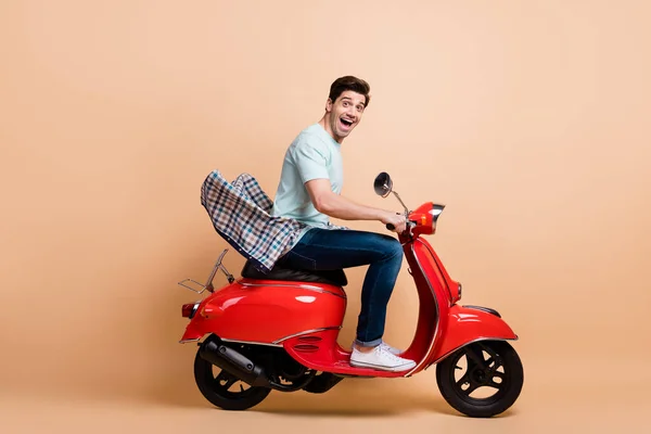 Profil oldalnézet portré a szép funky vidám srác élvezi vezetés piros moped gyors sebesség nap túra elszigetelt bézs pasztell szín háttér — Stock Fotó