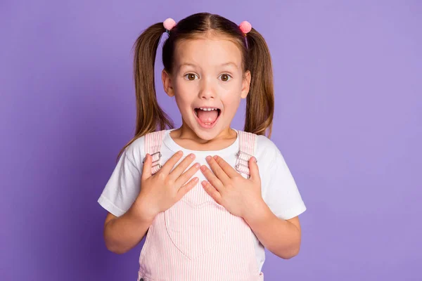 Photo of amazed cute little girl gesture show ask me wear pink overall dress isolated on violet color background