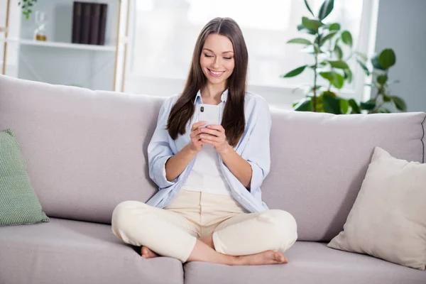 Plná velikost fotografie pěkné optimistické brunetky dlouhé účes dáma sedět na kouč vzhled telefon nosit modré tričko doma — Stock fotografie
