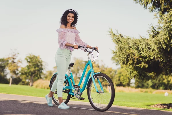 Foto de tamaño completo de la señora morena optimista de pie bicicleta usar pantalones de top de color lila zapatillas de deporte fuera en el parque —  Fotos de Stock