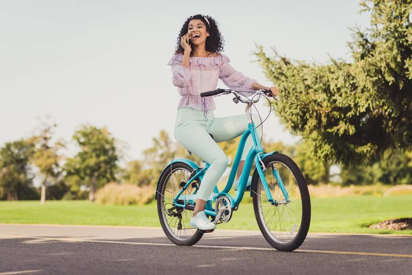 Foto de tamaño completo de la señora morena impresionada paseo bicicleta hablar teléfono usar pantalones superiores de color lila zapatillas de deporte fuera en el parque —  Fotos de Stock