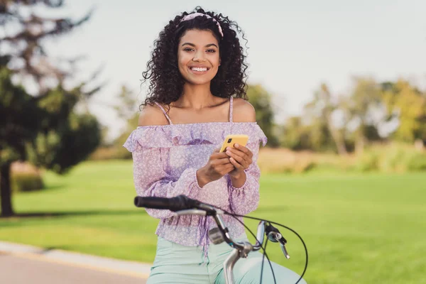 Foto de persona alegre y atractiva de piel oscura sentarse en bicicleta sostener teléfono mirar cámara dentada sonrisa tiempo libre al aire libre —  Fotos de Stock