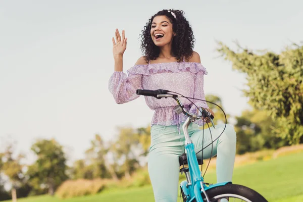 Retrato de excitado positivo escuro pele senhora sentar na bicicleta olhar interessado longe braço palma onda bom humor passeios ao ar livre — Fotografia de Stock