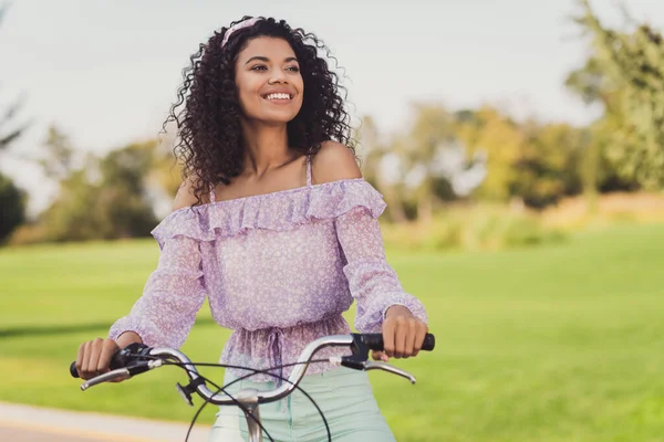 Foto de chica positiva de piel oscura sentarse en bicicleta mirar interesado lejos agradable fin de semana libre aventura al aire libre —  Fotos de Stock