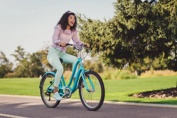 Foto de tamaño completo de la señora morena impresionada paseo en bicicleta usar pantalones superiores de color lila zapatillas de deporte fuera en el parque —  Fotos de Stock