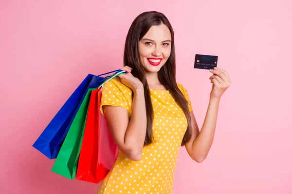 Фото счастливой красивой девушки носить желтые пунктирные футболки держать торговые сумки показывают кредитную карту изолированы на розовом фоне — стоковое фото