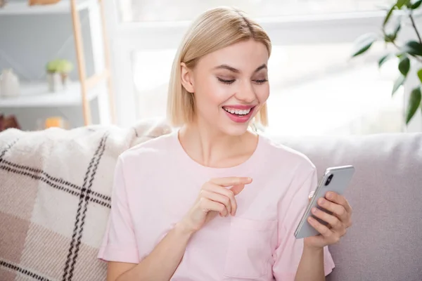 Фото веселой молодой блондинки держать за руки телефон улыбка сидеть диван в помещении дома — стоковое фото