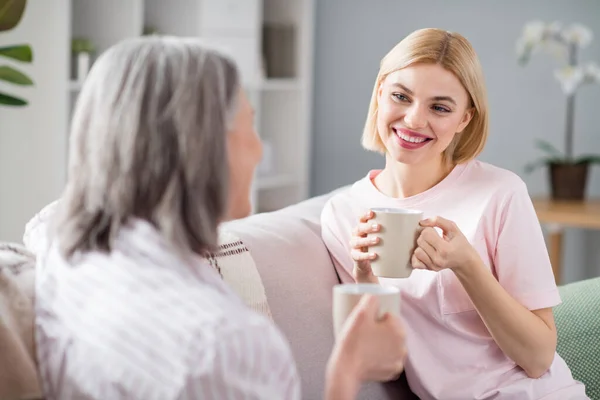 二人の正の女性の肖像はソファの上に座ってコーヒーを保持見てお互いに話す自由時間室内で通信 — ストック写真