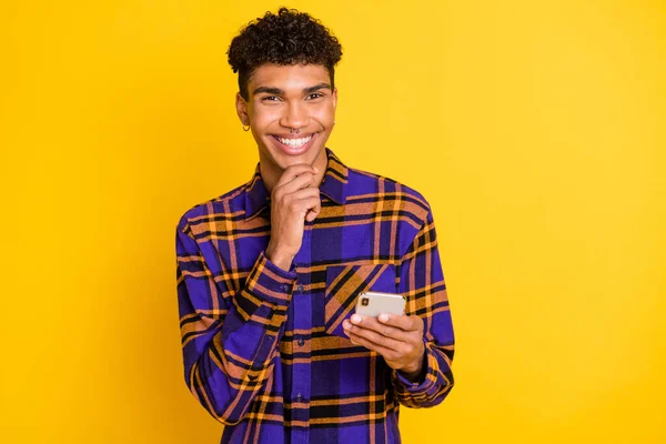 Foto van jonge afro guy gelukkig positieve glimlach hand raken kin denk bladeren mobiele telefoon geïsoleerd over gele kleur achtergrond — Stockfoto