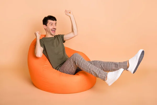 Foto van man sit beanbag raise vuisten look lege ruimte dragen groene t-shirt broek sneakers geïsoleerde beige kleur achtergrond — Stockfoto