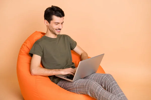 Foto von Kerl sitzen Sitzsack halten Netbook-Typisierung tragen grüne T-Shirt Hose isoliert beige Farbe Hintergrund — Stockfoto