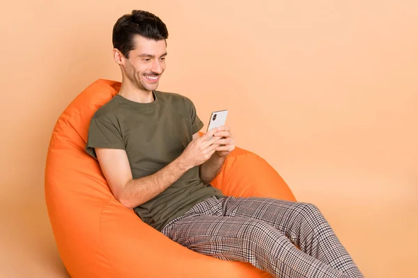 Foto retrato de chico usando aplicación en el teléfono inteligente sentado en silla naranja aislado sobre fondo de color beige pastel —  Fotos de Stock