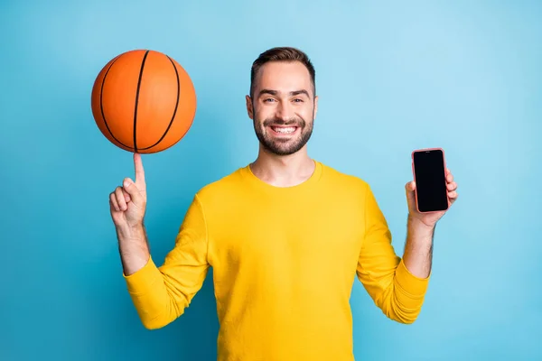 Foto van jong knap positief goed humeur lachende man houden telefoon en basketbal op vinger geïsoleerd op blauwe kleur achtergrond — Stockfoto