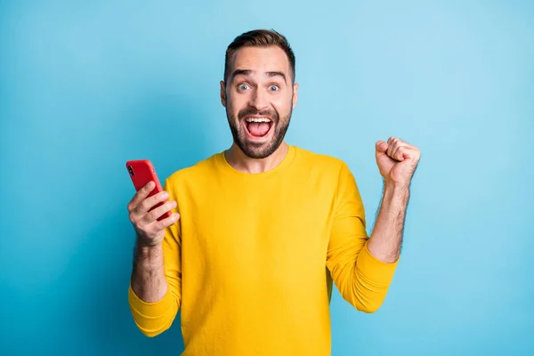 Foto van jong knap opgewonden gek glimlachen man verhogen vuist in de overwinning houden mobiele telefoon geïsoleerd op blauwe kleur achtergrond — Stockfoto