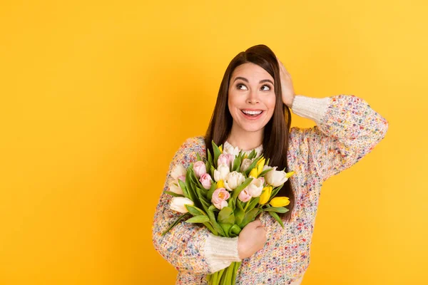 Fotografie šokované snivé roztomilé mladé ženy vypadají prázdné místo držet ruku hlavu květiny tulipány izolované na žlutém pozadí — Stock fotografie
