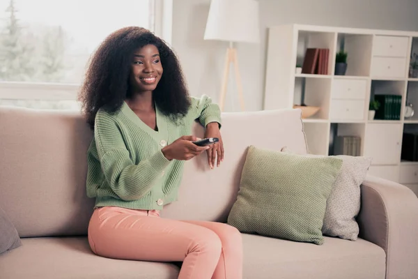 Foto de la joven atractiva mujer africana feliz sonrisa positiva sentarse sofá casa ver tv interruptor canal mando a distancia — Foto de Stock