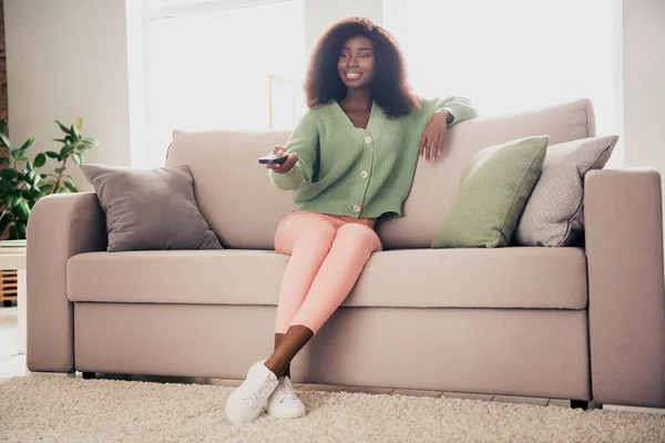 Retrato de chica atractiva de pelo ondulado alegre sentada en diván viendo series de televisión descansando en la casa plana sala de estar interior — Foto de Stock