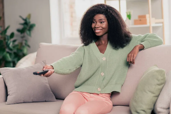 Retrato fotográfico de una chica rizada sonriente viendo la televisión sentada en el sofá en casa manteniendo el mando a distancia — Foto de Stock