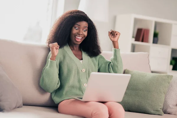 Retrato fotográfico de una chica rizada sonriente sentada en un sofá en casa manteniendo a la computadora portátil haciendo gestos como una ganadora riéndose de alegría — Foto de Stock