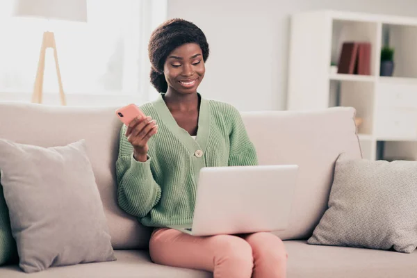Foto de joven atractiva mujer negra sonrisa feliz mirada portátil programador escribiendo reloj webinar uso teléfono celular sentarse sofá casa — Foto de Stock