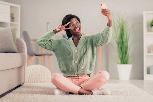 Genç heyecanlı afro kızın tam vücut fotoğrafı mutlu pozitif gülümseme selfie akıllı telefonunun huzur verici V-işareti evinde oturmasını sağlar. — Stok fotoğraf
