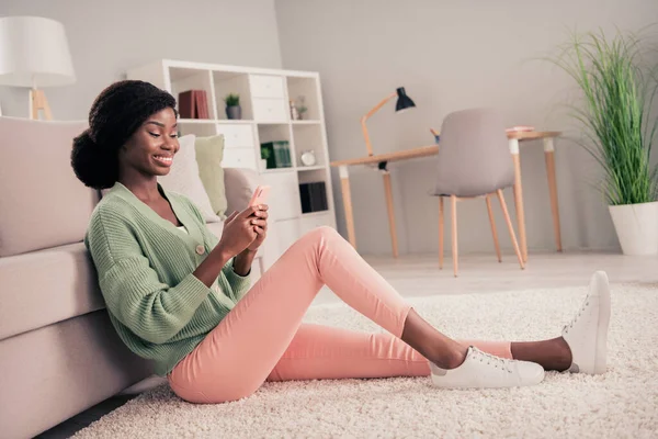 Foto lateral de perfil de longitud completa de la joven mujer negra feliz sonrisa positiva leer navegar por el teléfono celular sentarse piso de casa — Foto de Stock