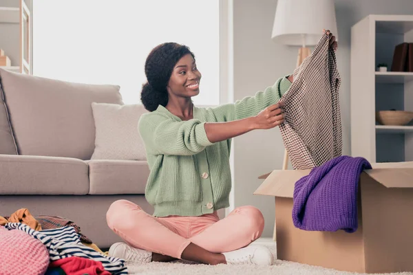 Retrato de chica alegre atractiva sentada en el suelo eligiendo ropa llevar tareas en casa plana sala de estar interior — Foto de Stock