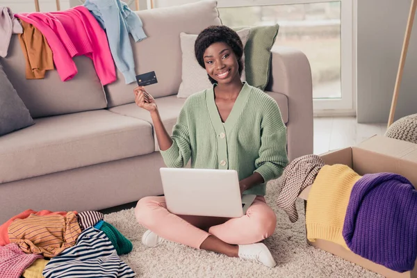 Longitud completa tamaño del cuerpo foto chica sentada en el suelo de mantenimiento de la computadora portátil que muestra la tarjeta bancaria sonriendo la venta de ropa — Foto de Stock