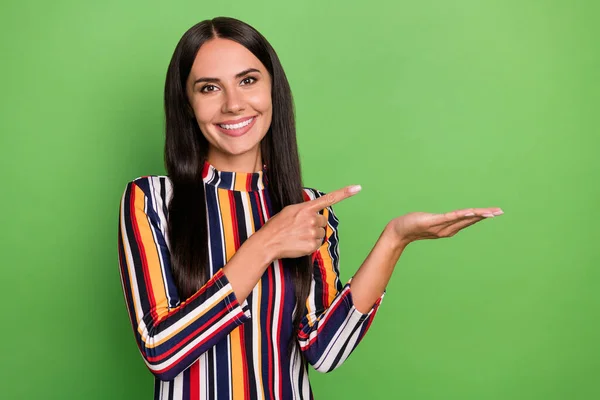 Fotografie optimistické brunetka dáma držet bod prázdný prostor nosit barevné pruhované tričko izolované na zeleném pozadí — Stock fotografie