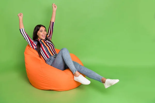 Potret gadis yang menarik dan ceria yang terbaring di kursi karung sambil bersenang-senang dengan mengangkat tangan terisolasi di atas latar belakang warna hijau cerah — Stok Foto